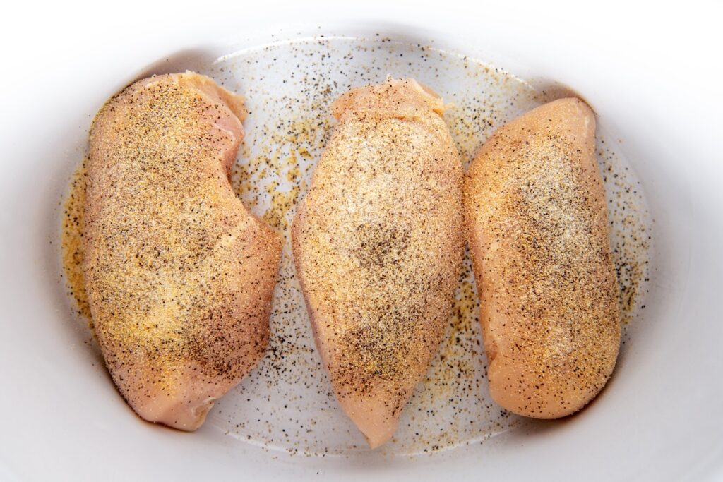 Three raw chicken breasts seasoned with black pepper and salt are arranged in a white baking dish.