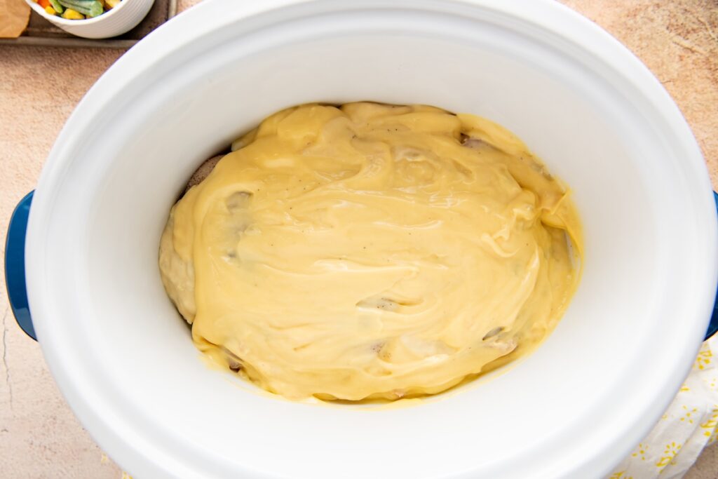 A slow cooker filled with a layer of creamy yellow sauce spread over raw chicken breasts.