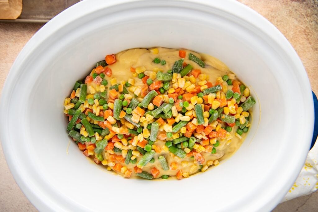 Frozen mixed vegetables and a creamy yellow sauce spread over chicken breasts in a white crockpot.