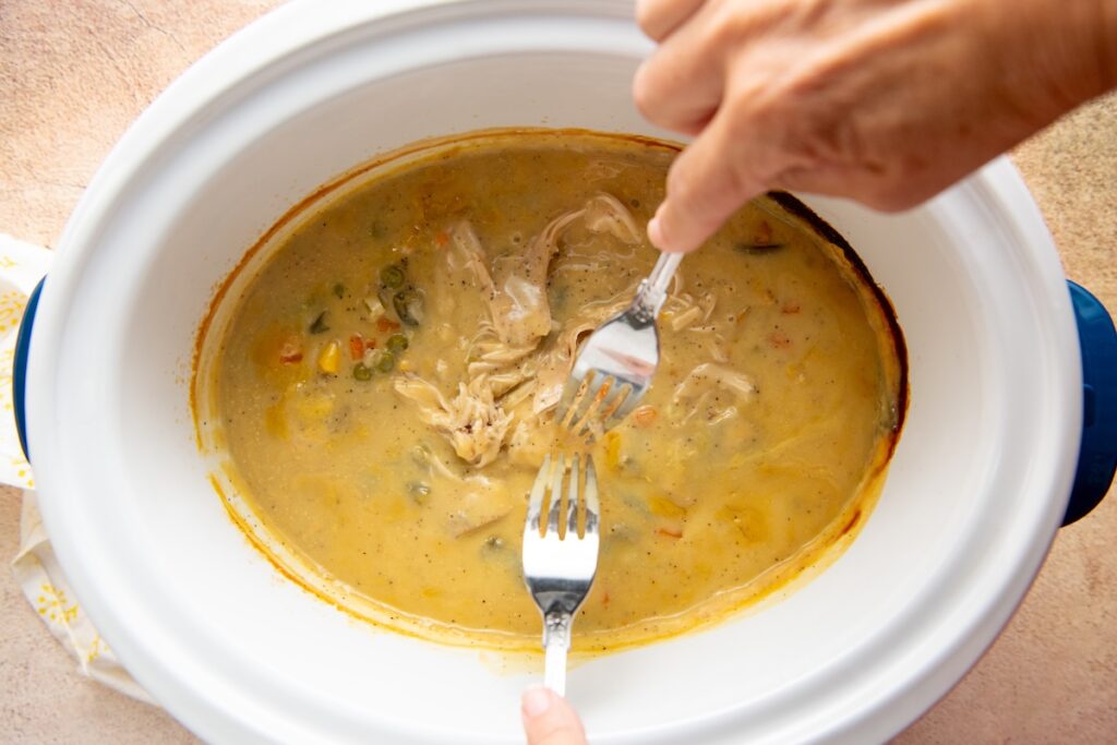 Two hands use forks to shred chicken in a creamy, yellow gravy with vegetables inside a white slow cooker.
