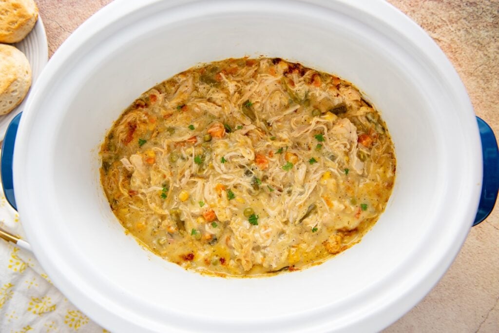 Crockpot chicken pot pie filling with peas, carrots, and corn in a creamy gravy inside a white crockpot, with biscuits on a plate nearby.