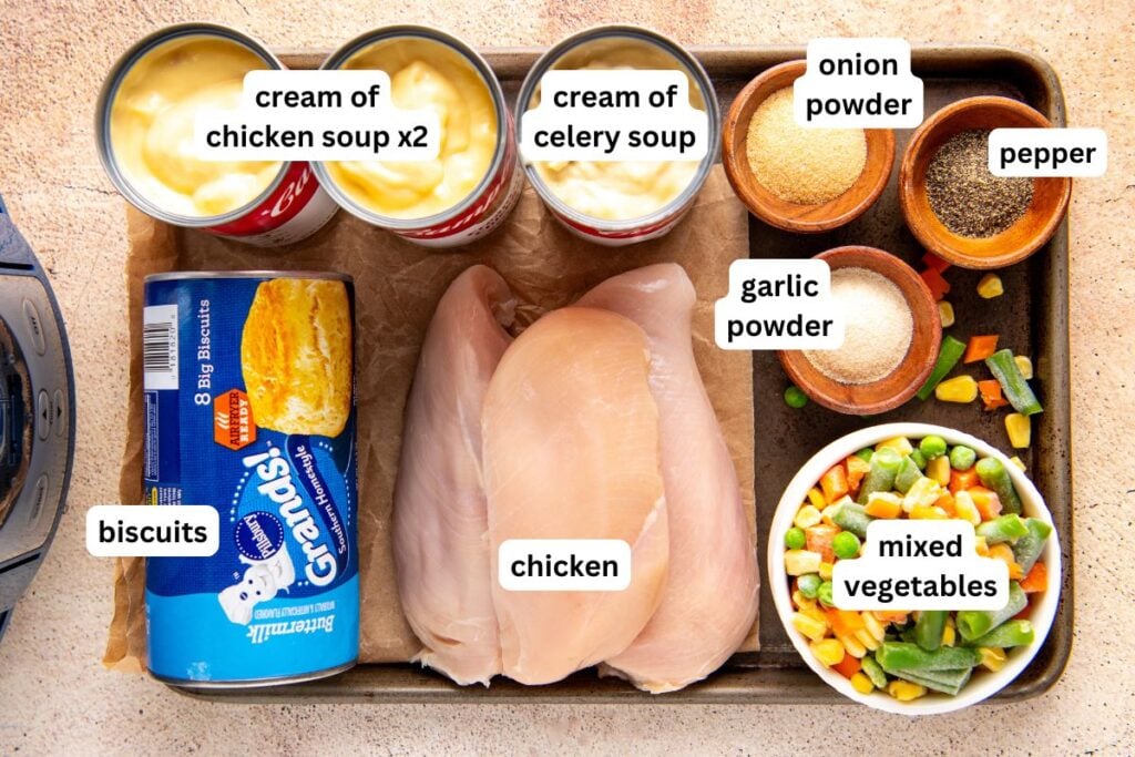 Dump-and-go Crockpot Chicken Pot Pie the whole family loves. Thick, creamy filling with chicken and veggies all served with a fluffy biscuit! Crock pot chicken pot pie recipe ingredients labeled and arranged on a tray: chicken breasts, canned cream of chicken and celery soups, canned biscuits, a bowl of mixed vegetables, and small bowls of onion powder, garlic powder, and pepper.