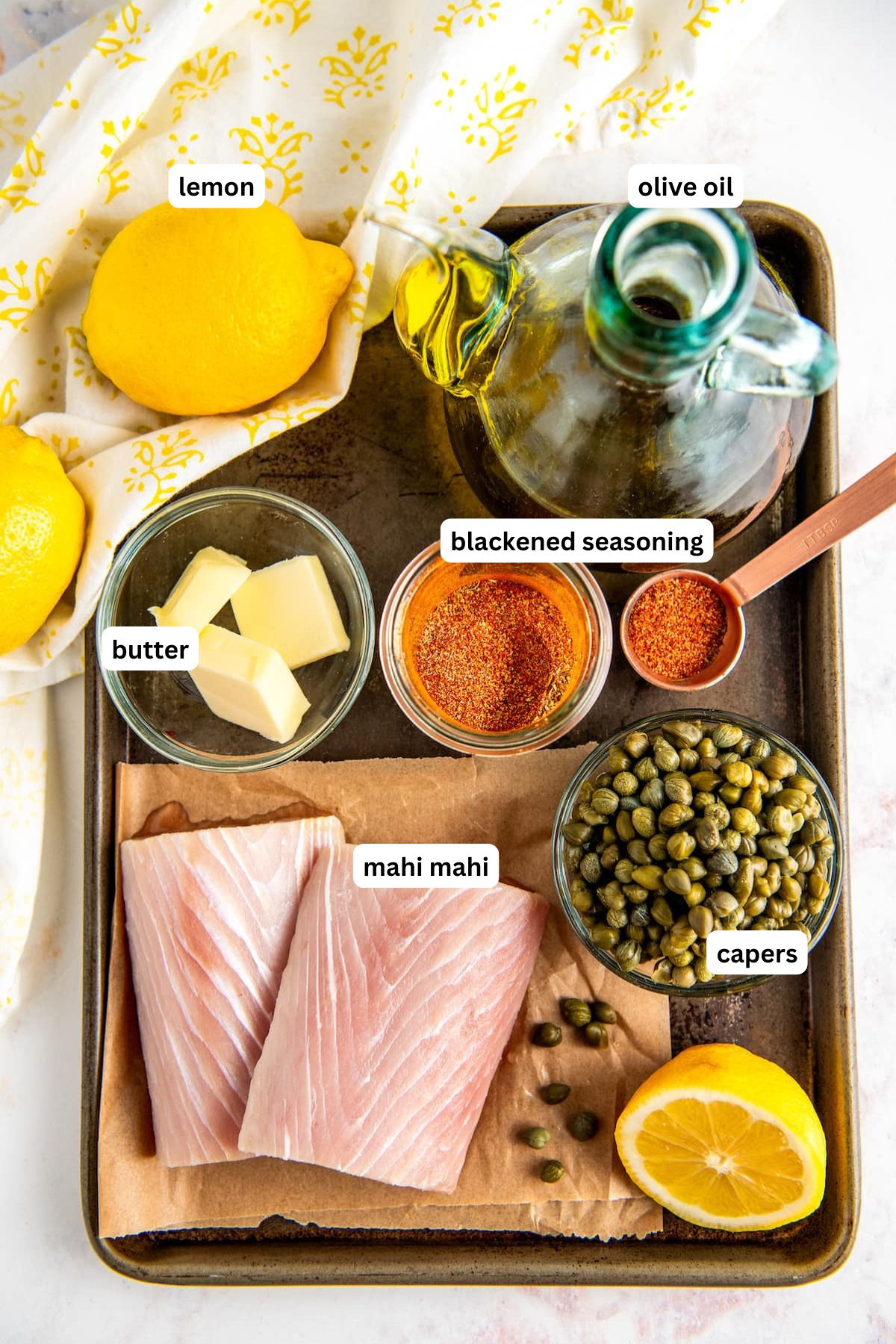 The ingredients for mahi mahi recipe are in order from top to bottom: lemon, olive oil, butter, cajun seasoning, mahi mahi, capers.