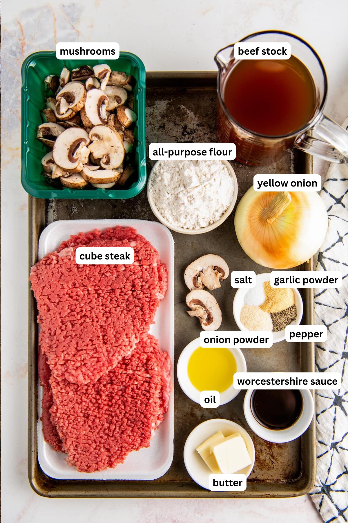 The ingredients for smothered cube steak recipe in order from top to bottom: mushrooms, beef stock, all purpose flour, yellow onion, cube steak, salt, garlic powder, onion powder, pepper, oil, worcestershire sauce, butter.