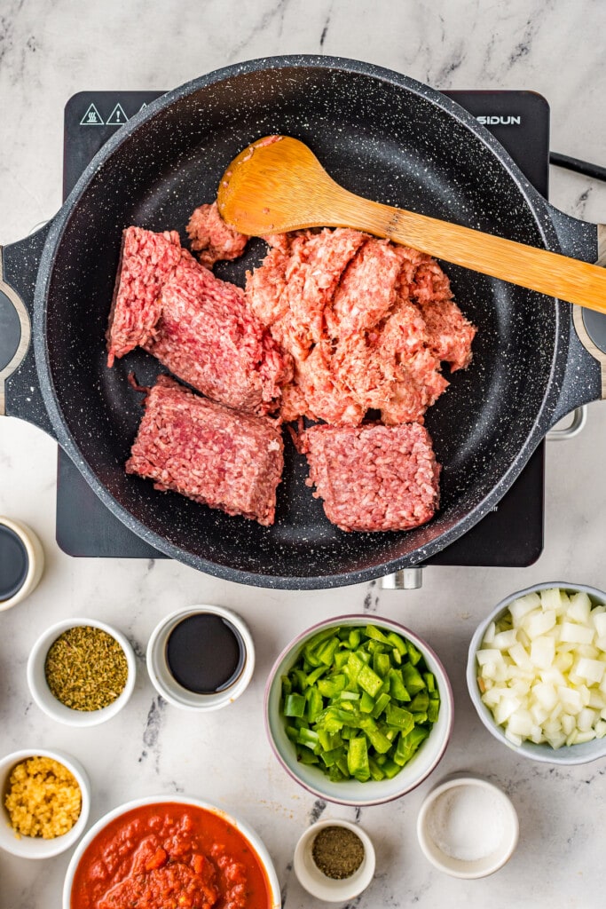 Overhead view of raw ground meat in a skillet with a wooden spoon, surrounded by bowls of veggies, spices, and tomatoes.