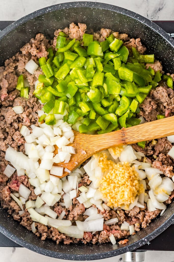 Overhead view of a skillet with cooked ground meat, topped with raw green bell peppers, onions, and garlic, with a wooden spoon.