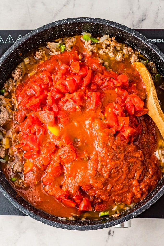 Overhead view of canned tomatoes and marinara sauce on top of cooked beef in a pot, with a wooden spoon.