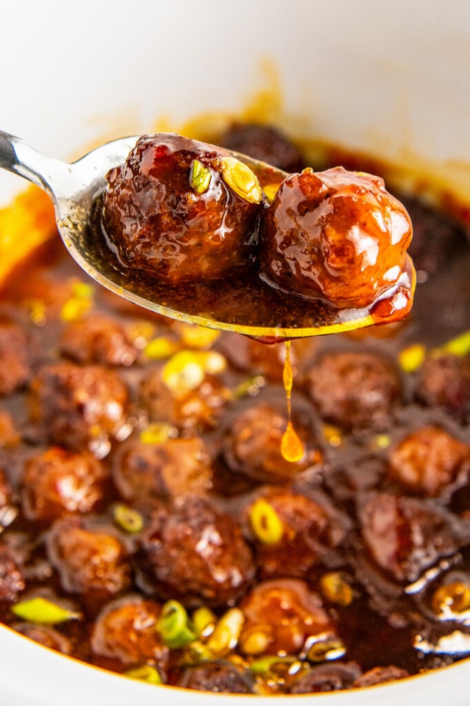 A spoon with two meatballs and sweet and sour sauce above a crockpot full of meatballs.