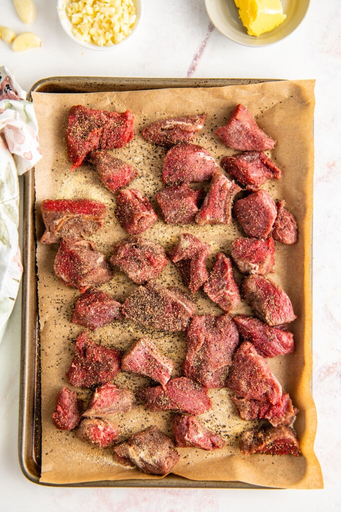 A bunch of seasoned raw bites of steak on a parchment paper-line baking sheat.