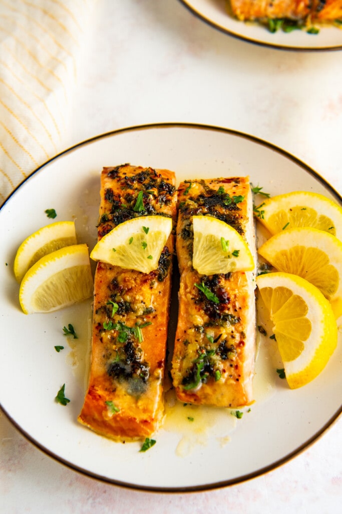 Overhead view of a plate with two salmon fillets topped with parsley and lemon, surrounded by lemon slices.