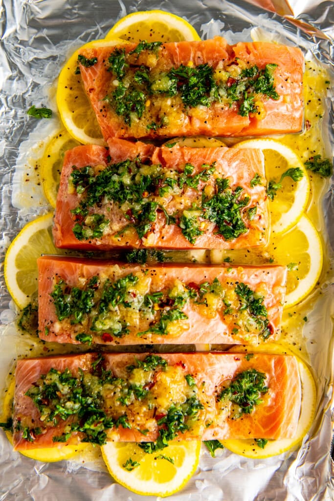 Overhead view of four raw salmon fillets on top of raw lemon slices, topped with an herb and garlic sauce.