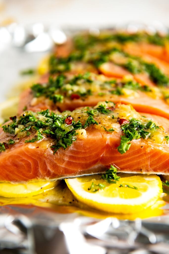 Side view of four raw salmon fillets topped with parsley and a sauce, on top of raw lemon slices.