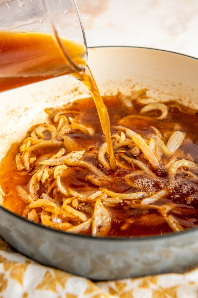 Chicken broth being poured into a pot of cooked onions.