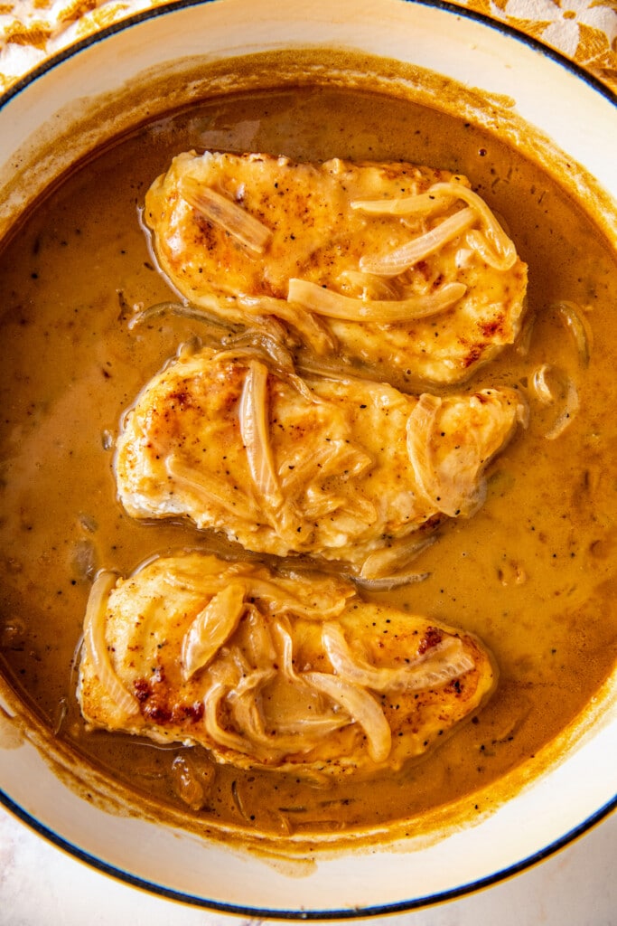 Overhead view of three chicken breasts cooking in a pot with gravy and onions.