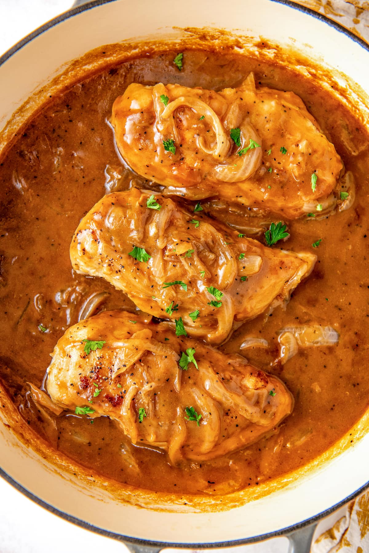 Overhead view of a pot filled with three smothered chicken breasts with onions in a skillet garnished with fresh parsley. 