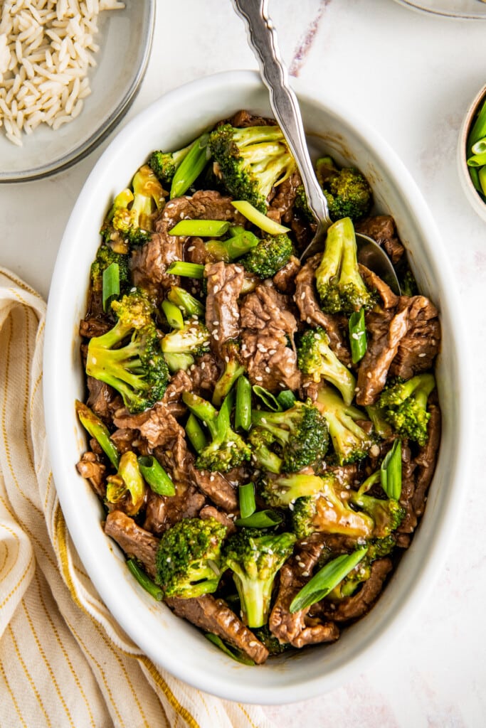 A white serving dish filled with beef and broccoli garnished with green onions and sesame seeds.