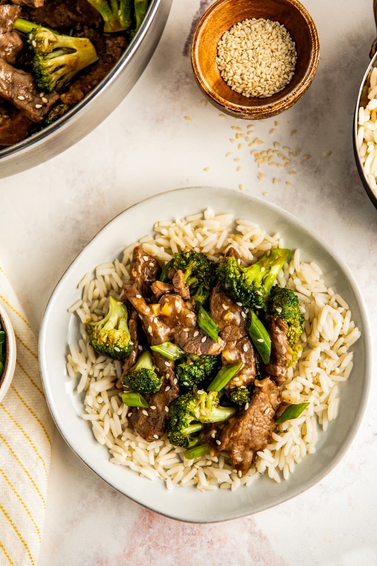 A bowl of beef and broccoli over white rice with a bowl of sesame seeds in the background.