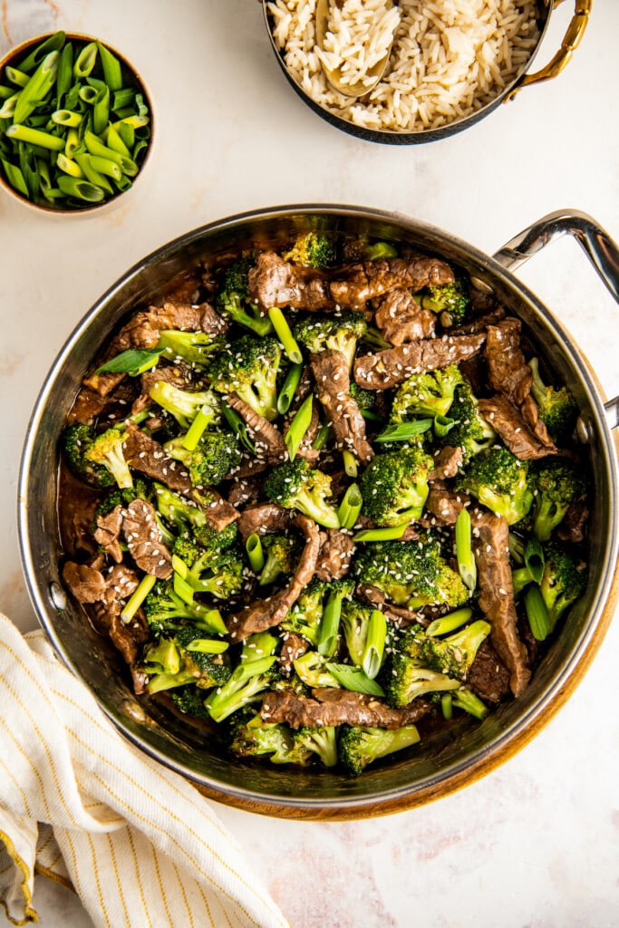 A pot of beef and broccoli on the counter with a bowl of white rice and green onions in the background.