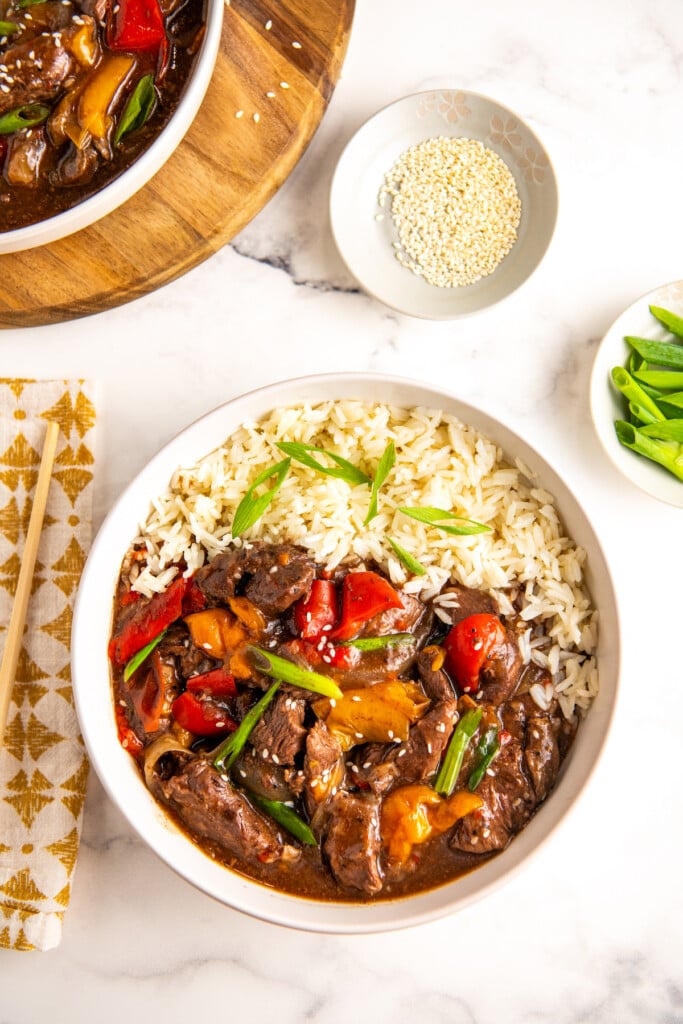 Crockpot pepper steak served in a white bowl over rice. Garnished with green onions and sesame seeds.