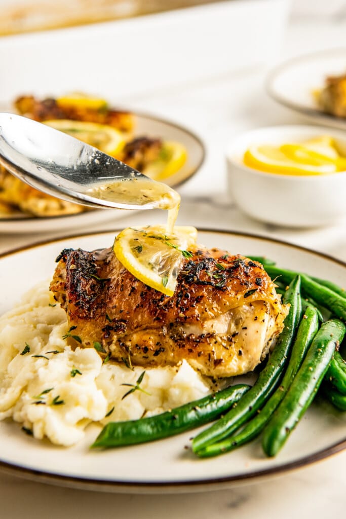 Spooning drippings over lemon pepper baked chicken on a plate with mashed potatoes and green beans.