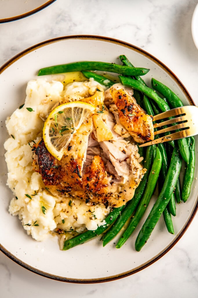 A fork taking a bite of a baked lemon pepper chicken thigh over a bed of mashed potatoes with a side of green beans.