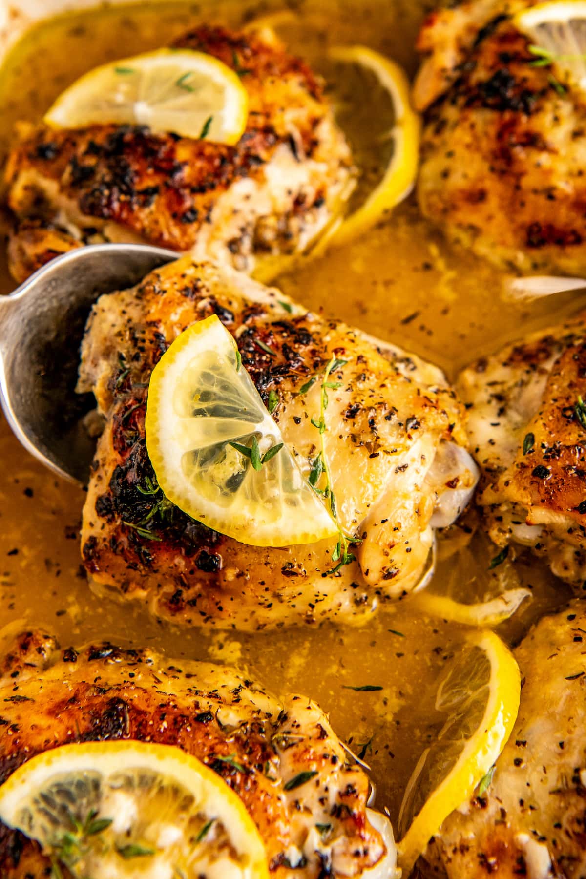 Close up of baked lemon pepper chicken in a casserole dish with a serving spoon lifting up a baked thigh.