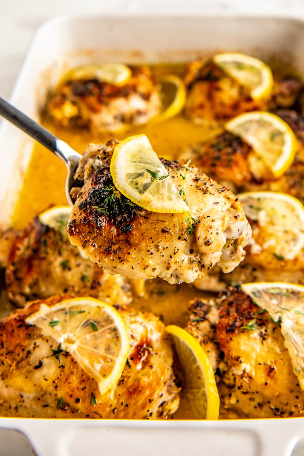 A serving spoon lifting a piece of lemon pepper baked chicken out of the roasting pan.