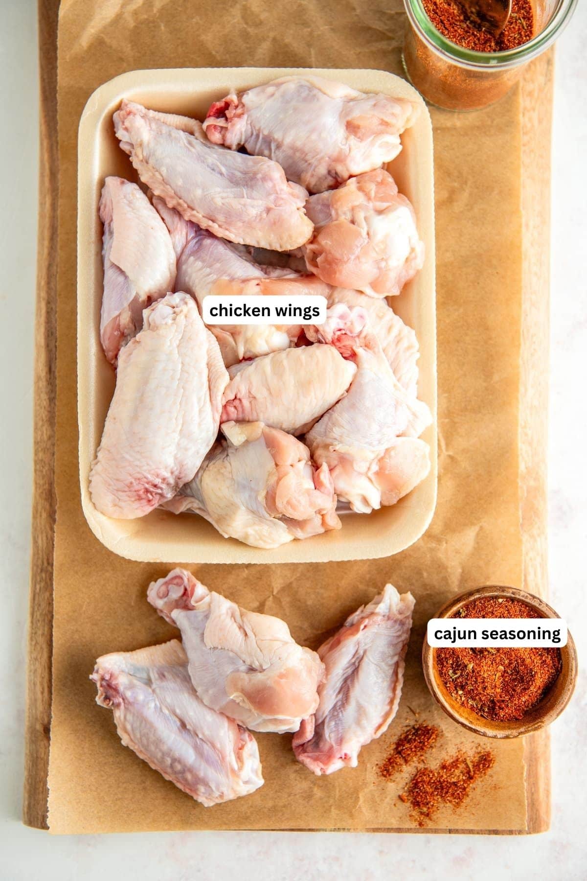 Ingredients for oven baked wings recipe arranged on a cutting board. From top to bottom: raw chicken wings and a bowl of cajun seasoning.