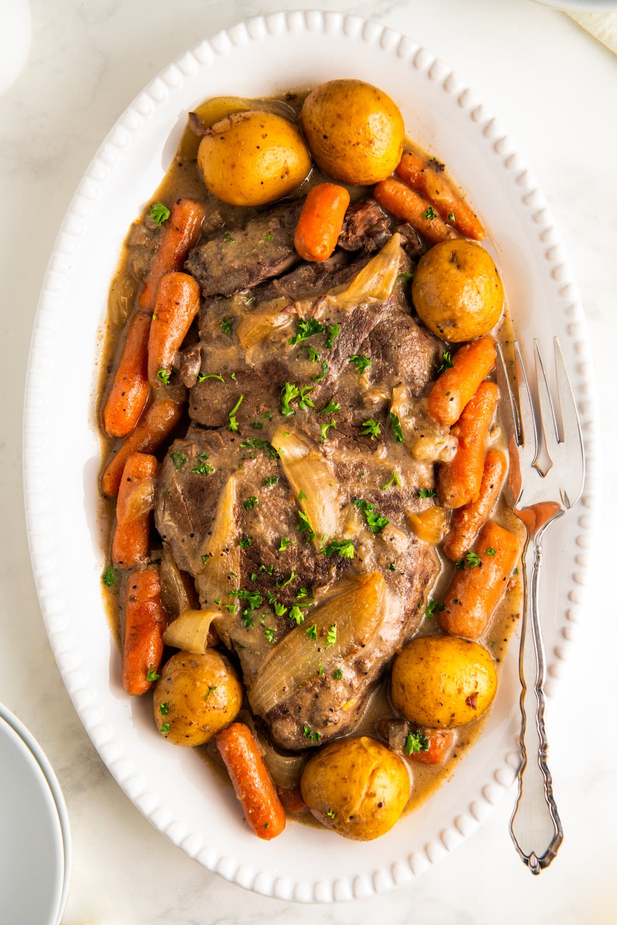 Crock pot London broil with vegetables in a brown gravy on a white serving platter with a serving fork on the side.