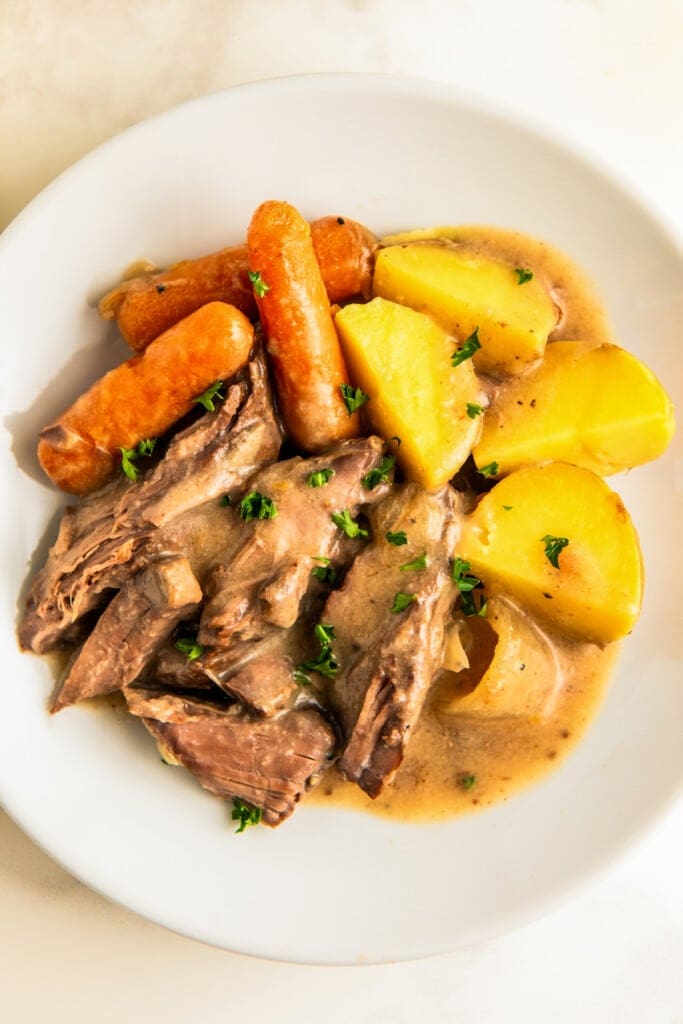 A plate with sliced slow cooker London broil, potatoes, onions and baby carrots covered in rich gravy.