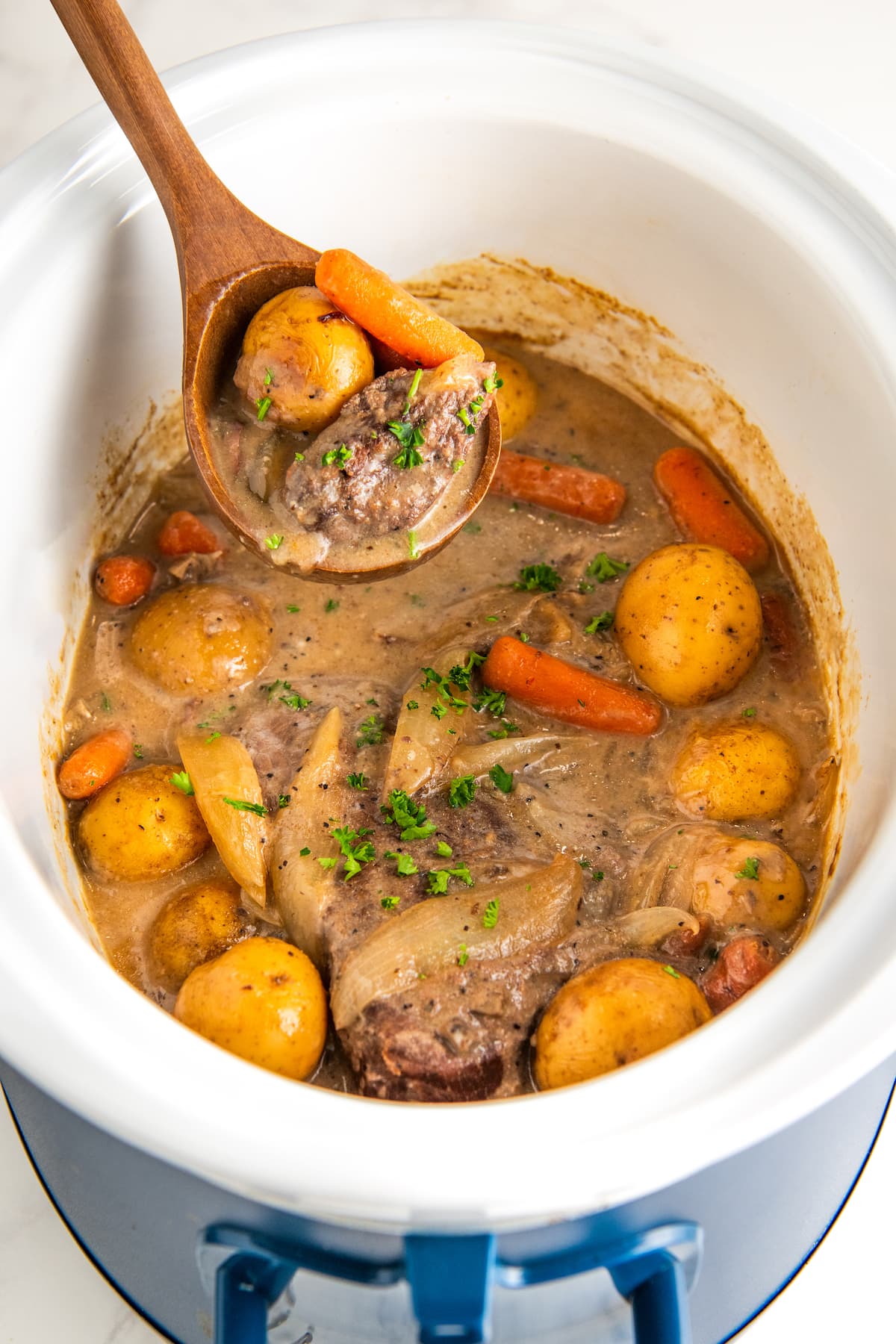 Tender London broil in crock pot cooked with vegetables in a brown gravy with a wooden serving spoon scooping out a serving.