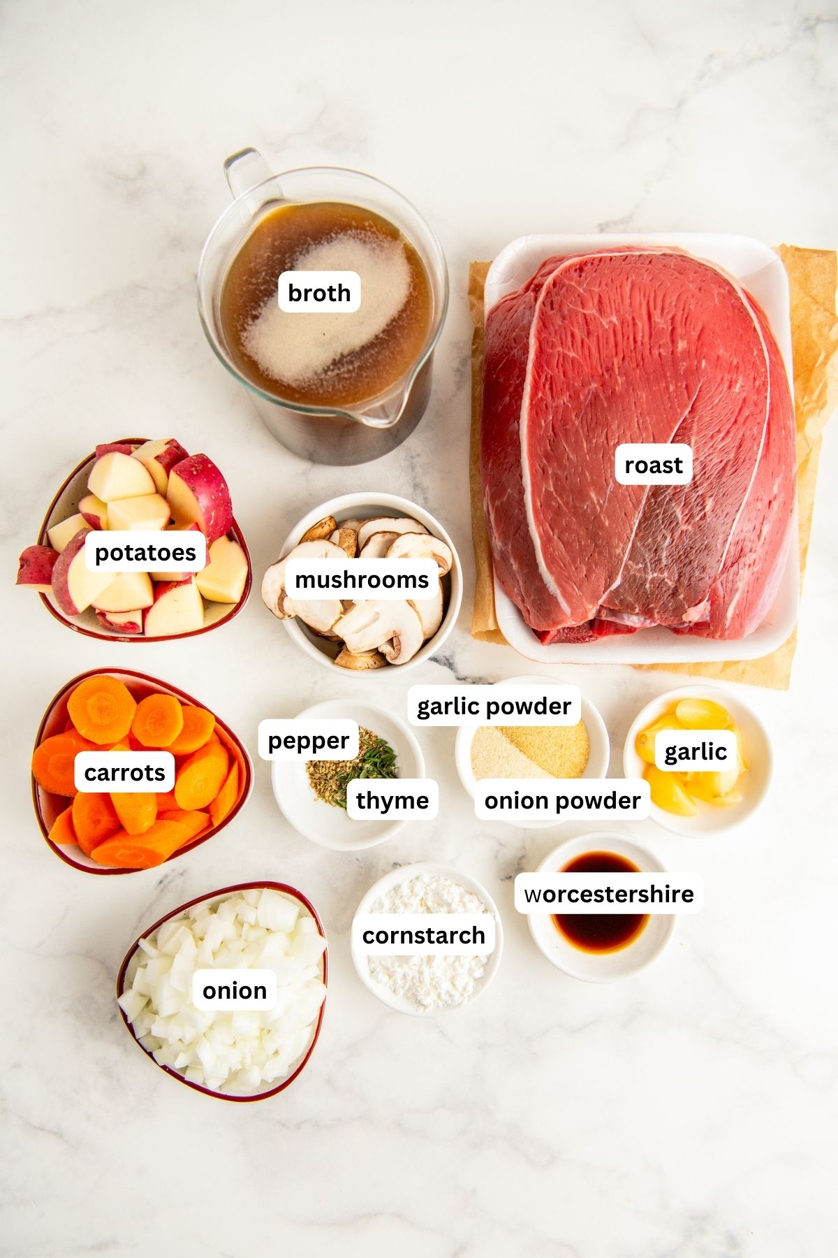 Ingredients for crockpot pot roast recipe arranged in bowls on a marble countertop, from top to bottom: broth, roast, potatoes, mushrooms, carrots, pepper, thyme, garlic powder, onion powder, garlic, onion, cornstarch and Worcestershire sauce.