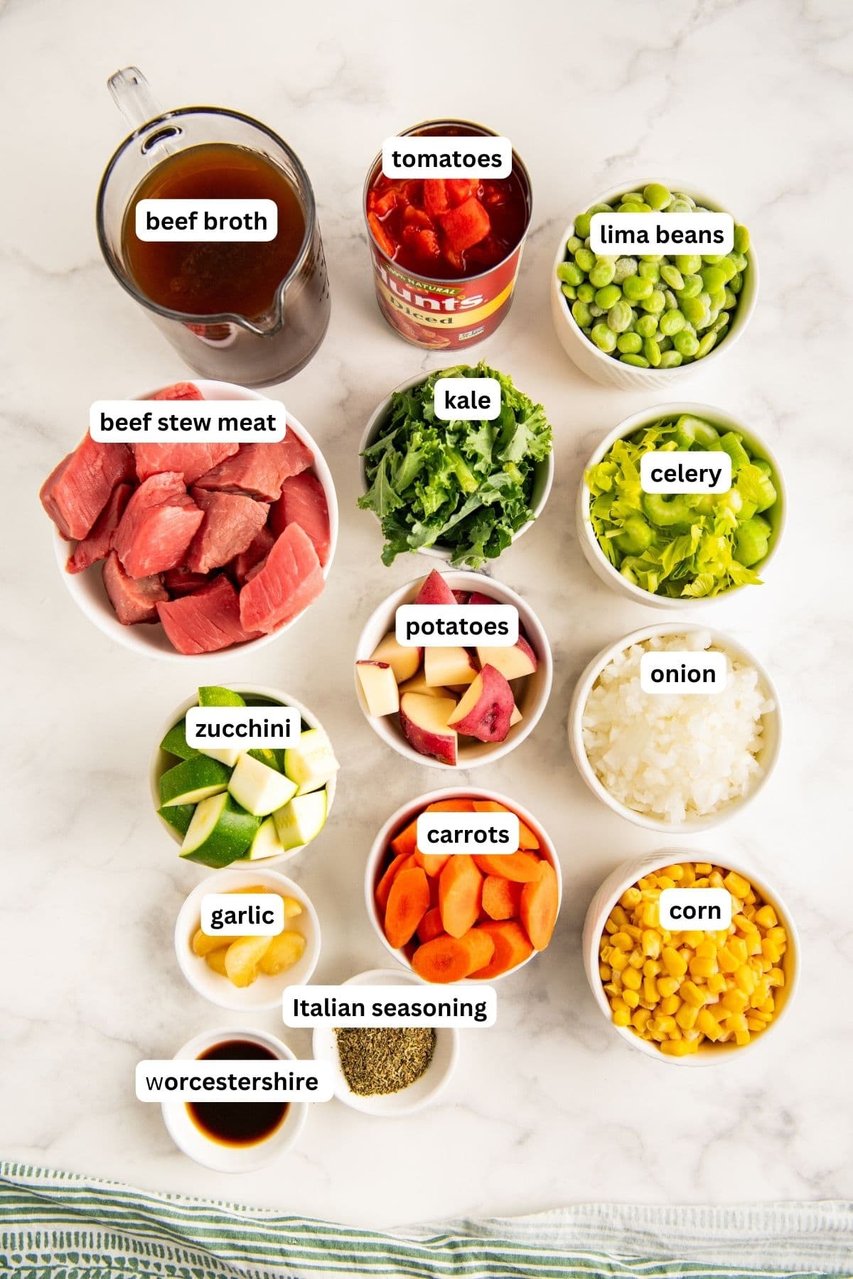 Ingredients for crockpot vegetable beef soup arranged in bowls on a countertop, from top to bottom: beef broth, tomatoes, lima beans, beef stew meat, kale, celery, potatoes, onion, zucchini, carrots, garlic, corn, Italian seasoning and Worcestershire sauce.