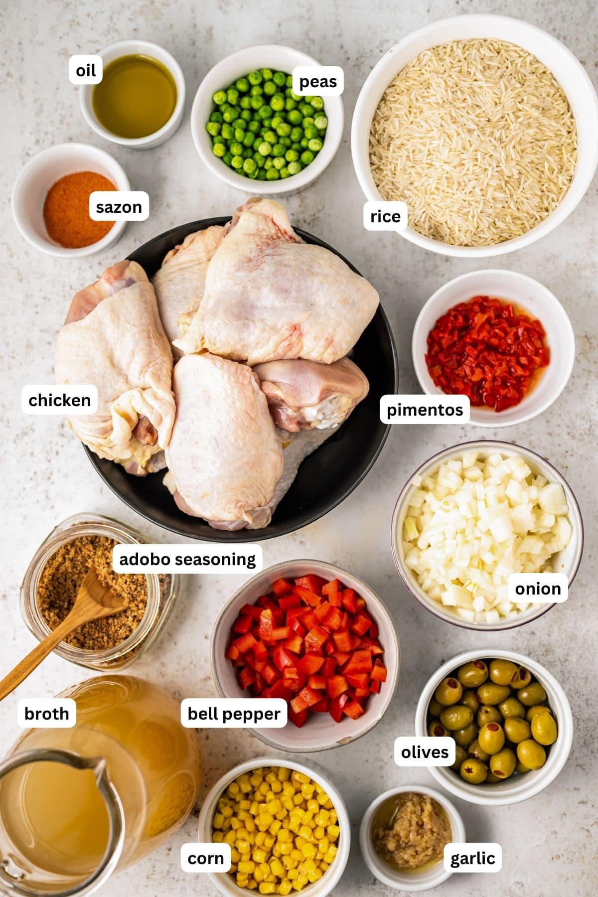 Ingredients for arroz con pollo recipe arranged in bowls, from top to bottom: oil, peas, rice, sazon, chicken, pimentos, onion, adobo seasoning, bell pepper, olives, broth, corn and garlic.