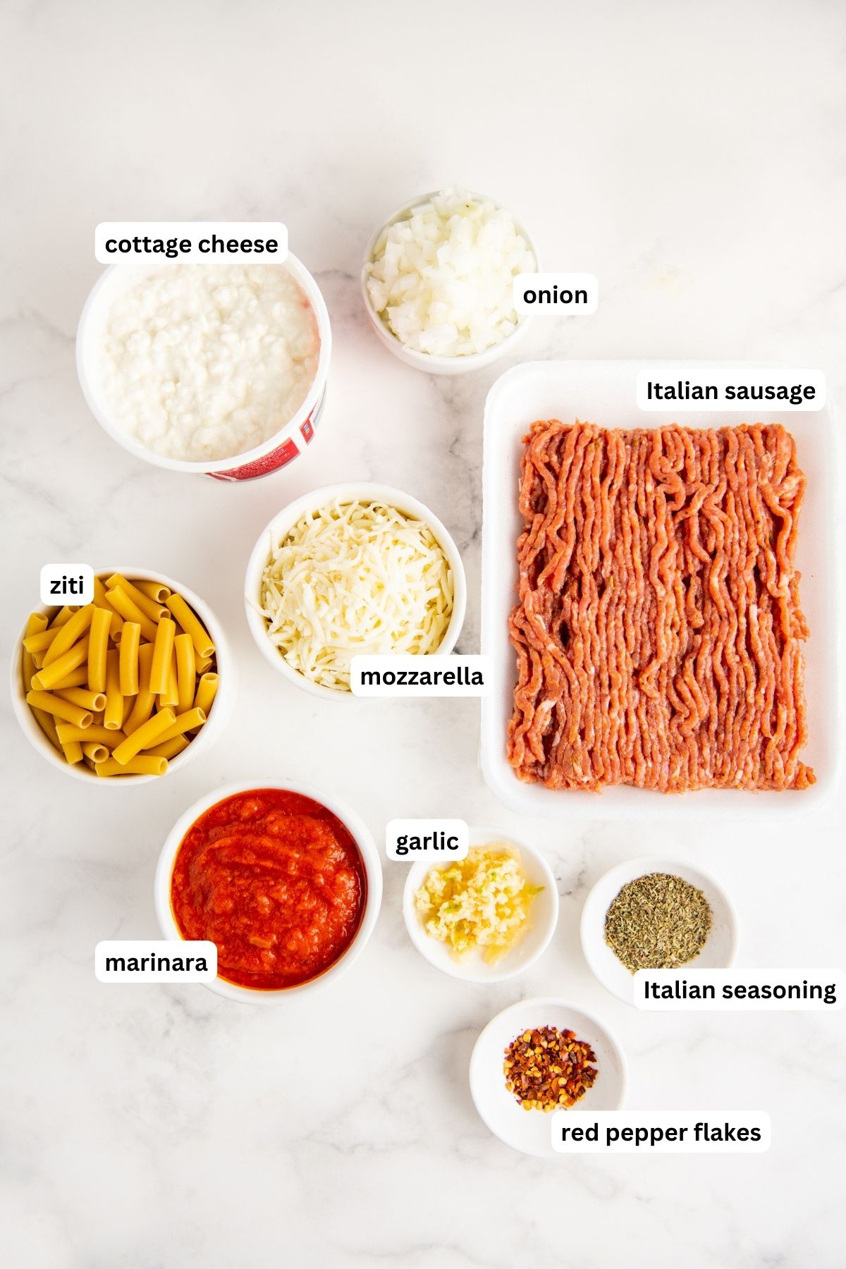 Baked ziti recipe ingredients arranged in bowls on a marble countertop, from top to bottom: onion, cottage cheese, Italian sausage, mozzarella, ziti pasta, marinara, garlic, Italian seasoning and crushed red pepper flakes.