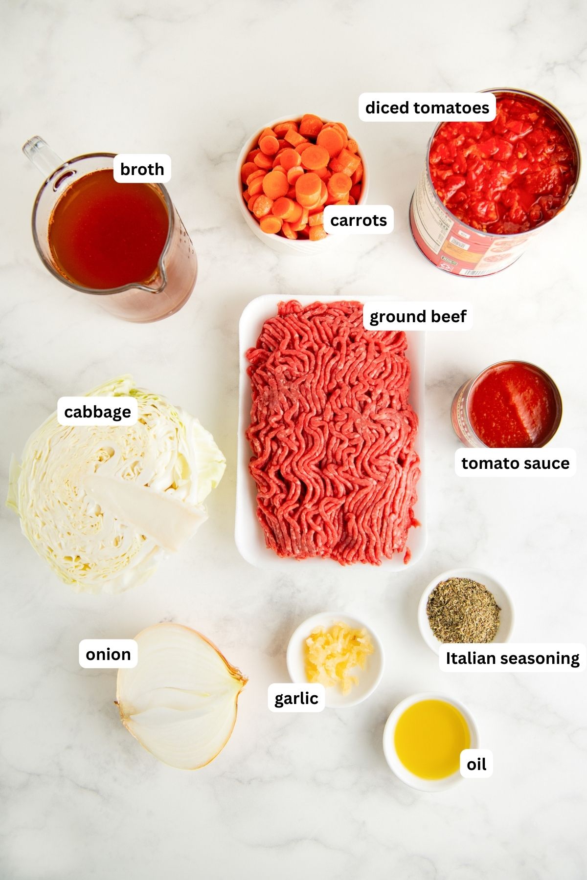 Ingredients for cabbage roll soup recipe arranged in bowls on a marble counter top. From top to bottom: diced tomatoes, carrots, broth, ground beef, cabbage, tomato sauce, onion, Italian seasoning, garlic and oil.