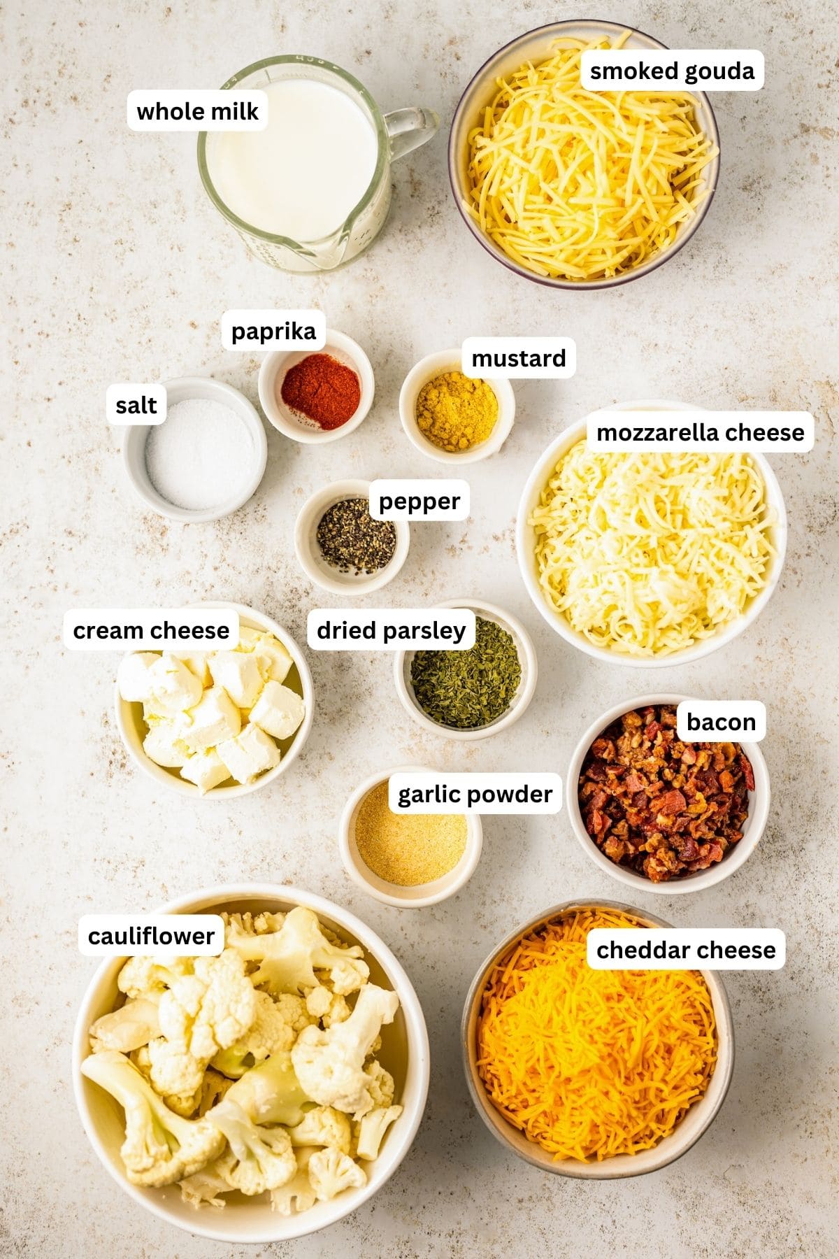 Ingredients for cauliflower Mac and cheese recipe arranged in bowls, from top to bottom: smoked gouda shredded cheese, whole milk, paprika, mustard, salt, shredded mozzarella cheese, cream cheese, dried parsley, bacon bits, garlic powder, cauliflower florets and shredded cheddar cheese.