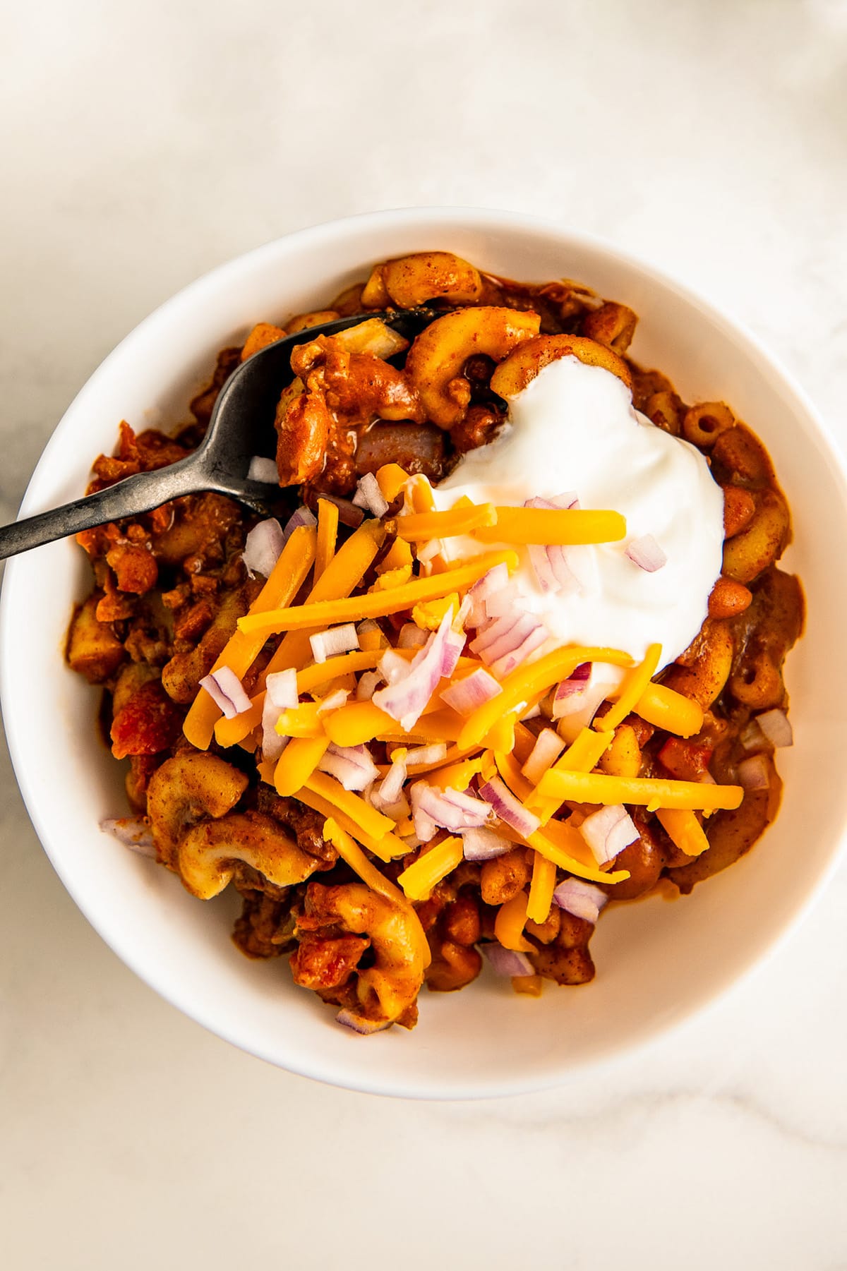 A bowl of mac and cheese chili topped with shredded cheese, sour cream and onions on top.