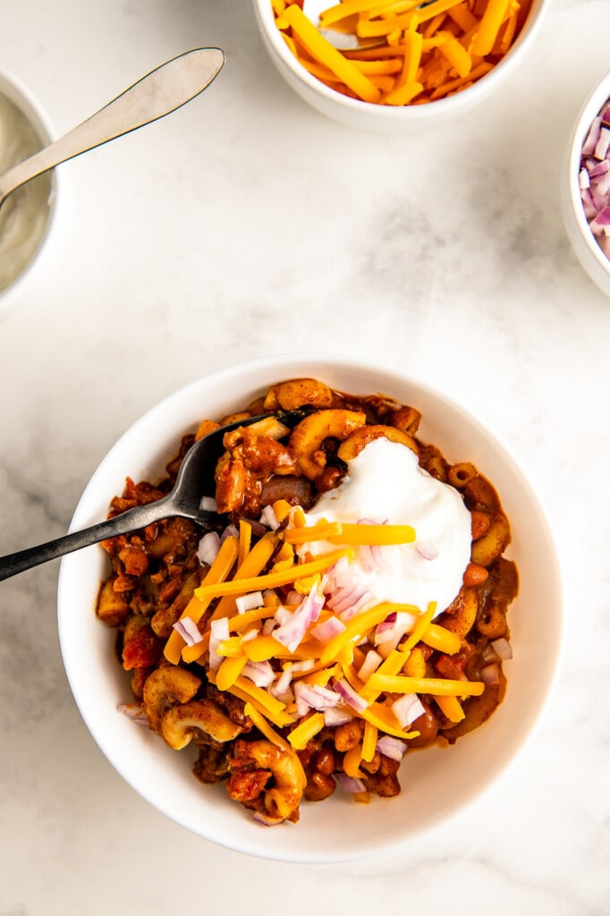A bowl of chili mac and cheese topped with sour cream, shredded cheese and diced onions with a spoon ready to take a bite.