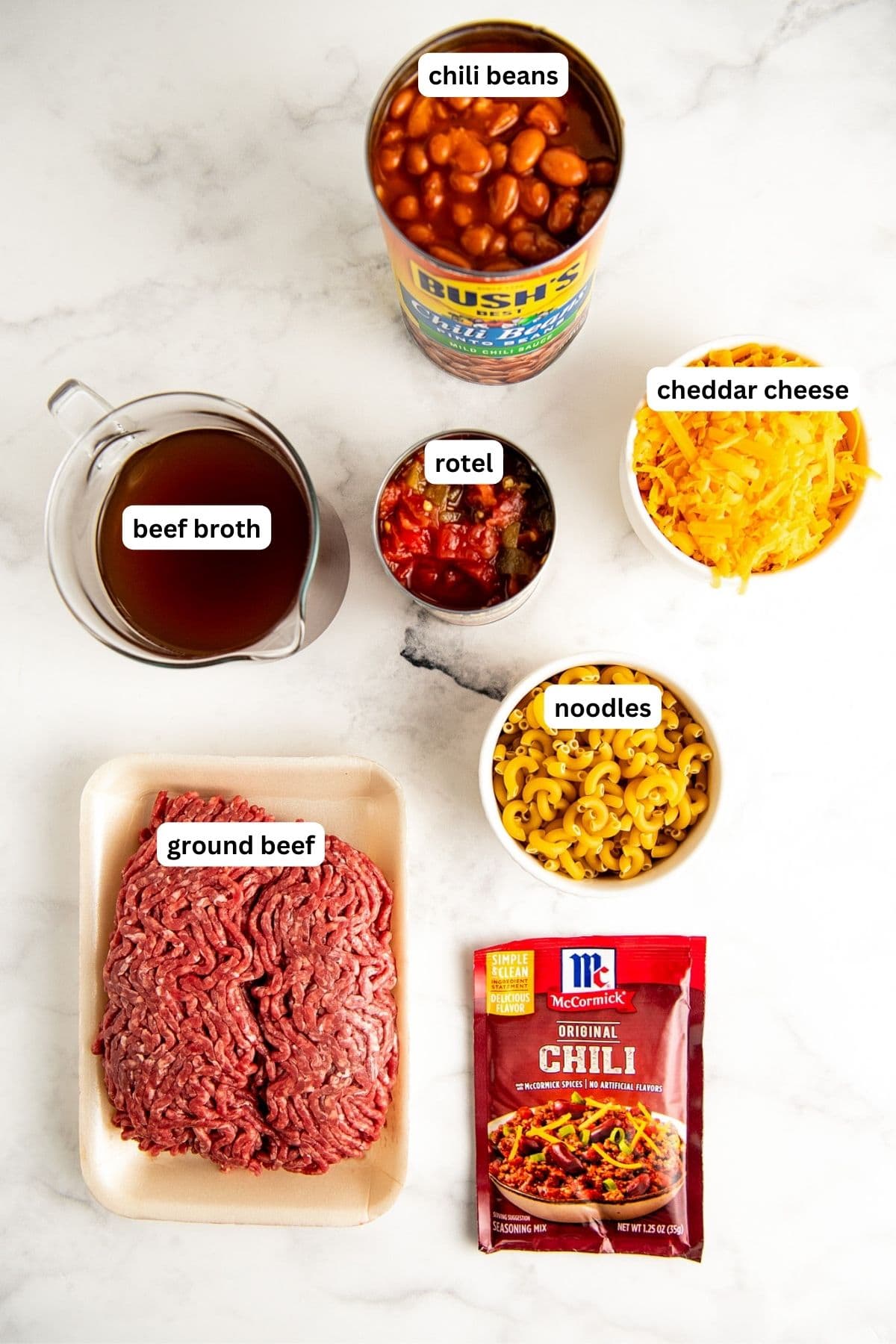 Ingredients to make crockpot chili mac & cheese recipe, from top to bottom: chili beans, cheddar cheese, rotel, beef broth, noodles, ground beef and chili seasoning.