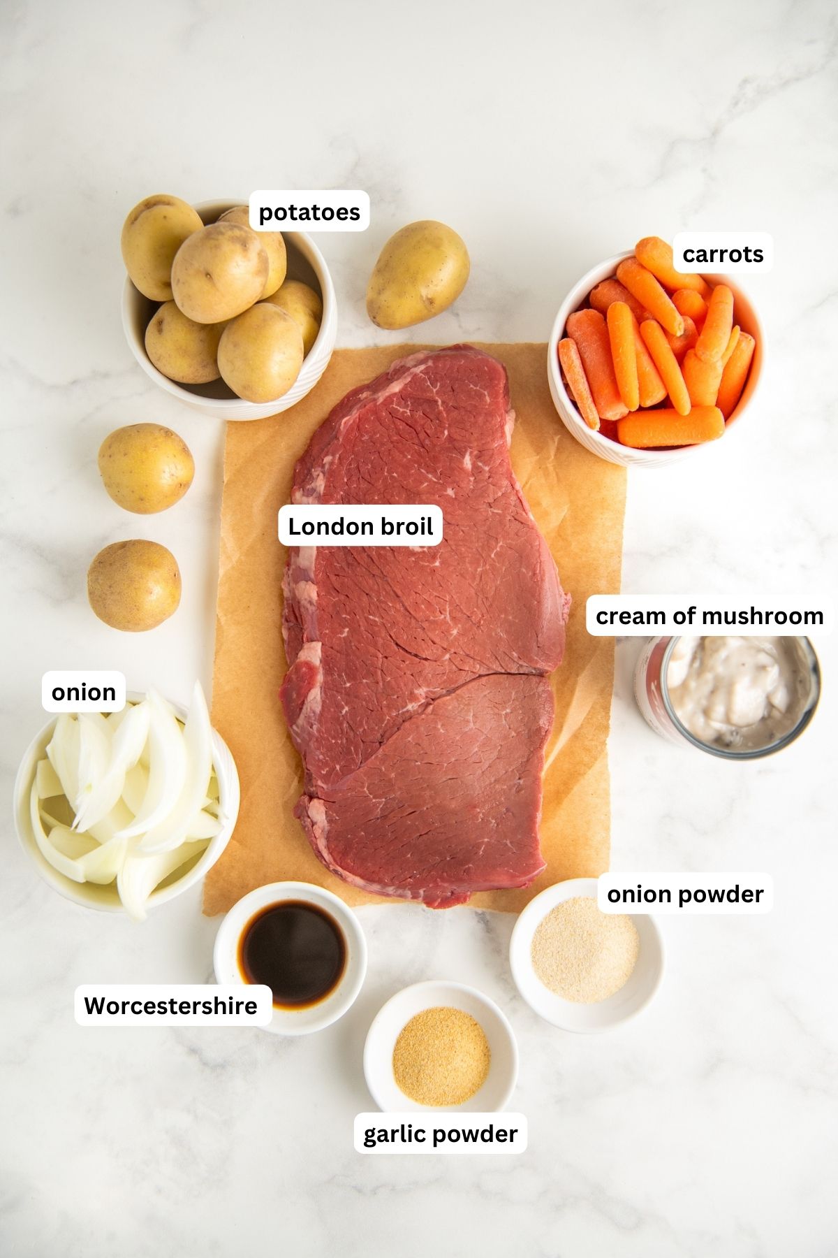 Ingredients for London broil in crockpot arranged in bowls on a marble countertop, from top to bottom: potatoes, carrots, London broil, onion, cream of mushroom, Worcestershire sauce, onion powder and garlic powder.