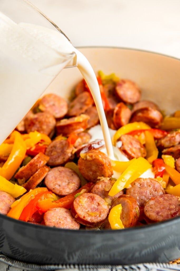 Adding heavy cream to a skillet filled with bell peppers and sliced smoked sausage. 