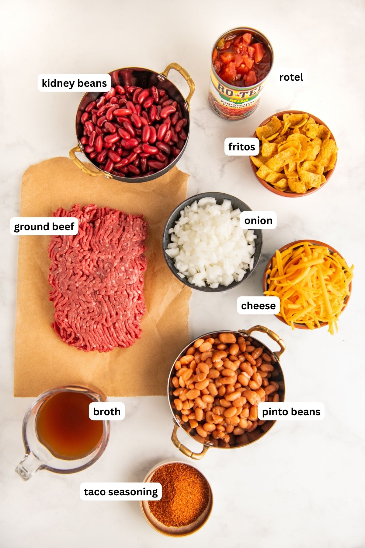 Ingredients to make Frito recipe arranged in bowls. From top to bottom: rotel, kidney beans, fritos, ground beef, onion, cheese, pinto beans, broth and taco seasoning.