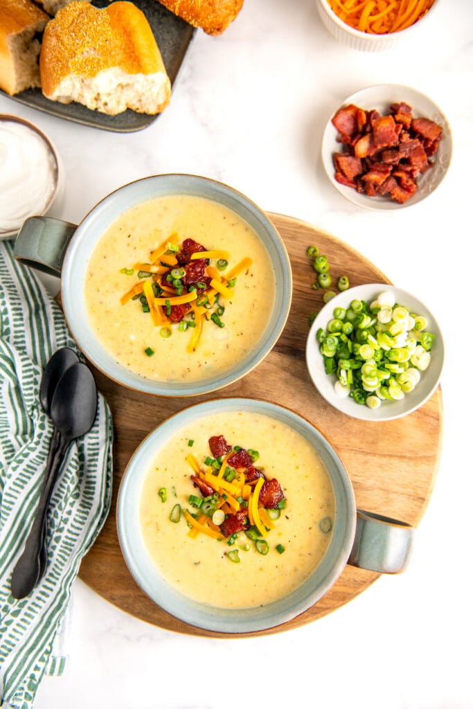 2 bowls of instant pot baked potato soup with garnishes and bread in the background.