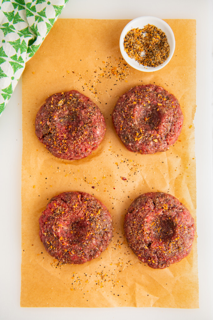 Four burger patties, each with an indent in the center, on a wooden cutting board next to a small bowl of seasoning.