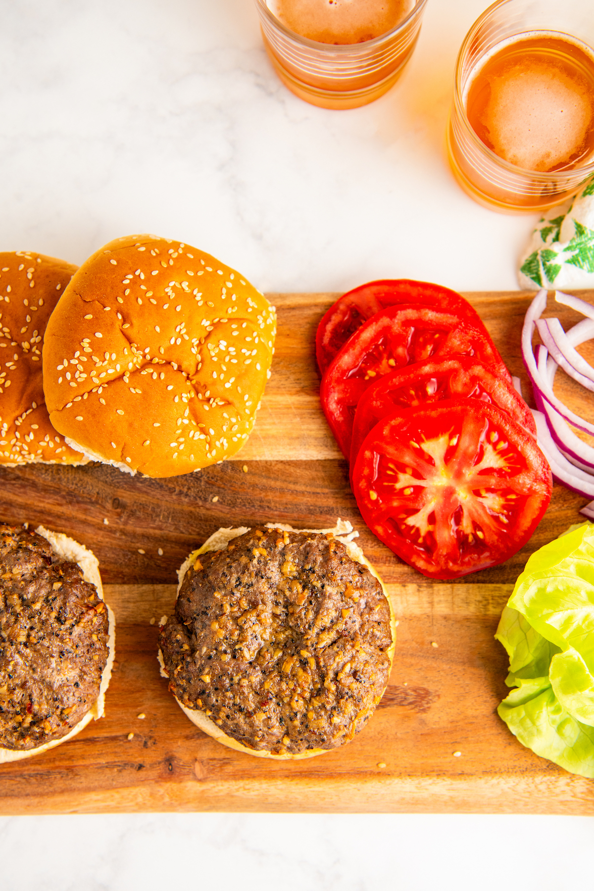 Overhead view of two partially assembled air fryer burgers on a wooden board, next to sliced tomatoes and onions, and two top buns.