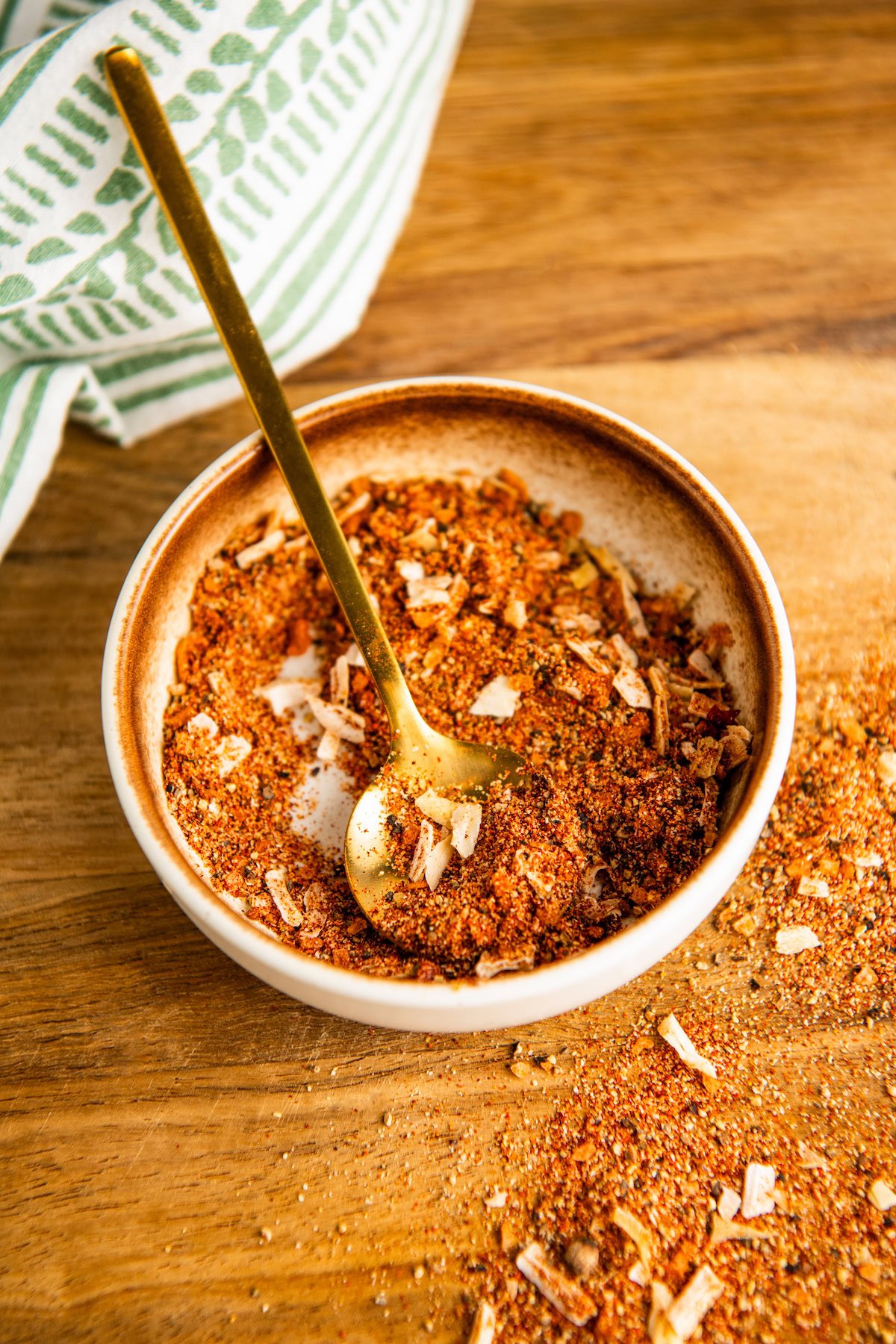 A golden spoon resting in a bowl of the best burger seasoning, on a wooden countertop.