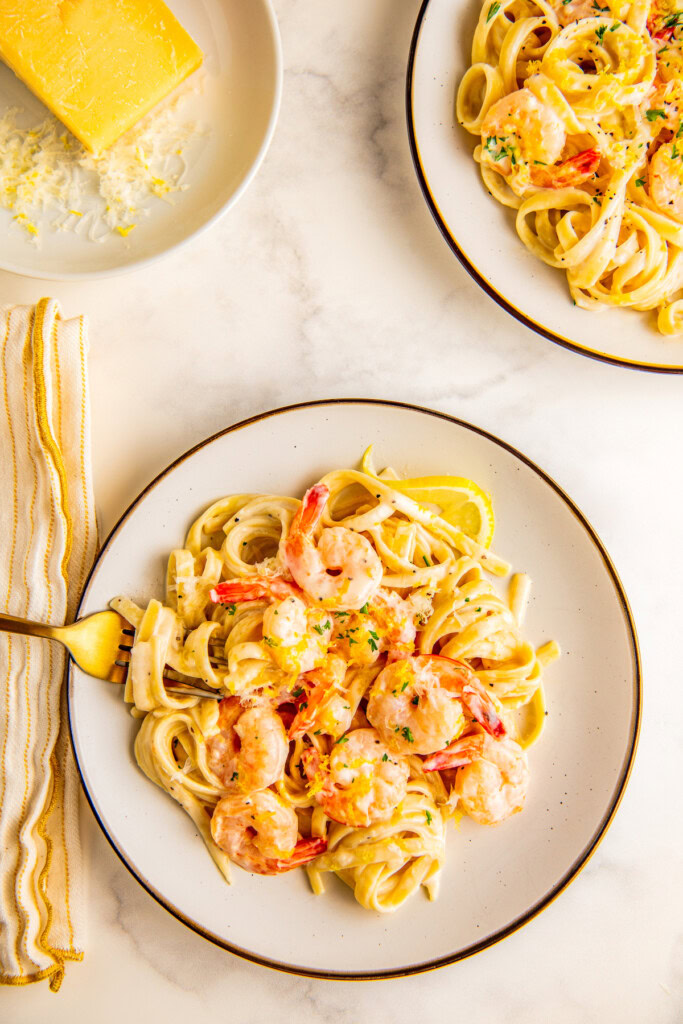 A plate of creamy pasta with shrimp lemon and garlic on a plate topped with fresh lemon zest and a bowl of grated parmesan cheese in the background.