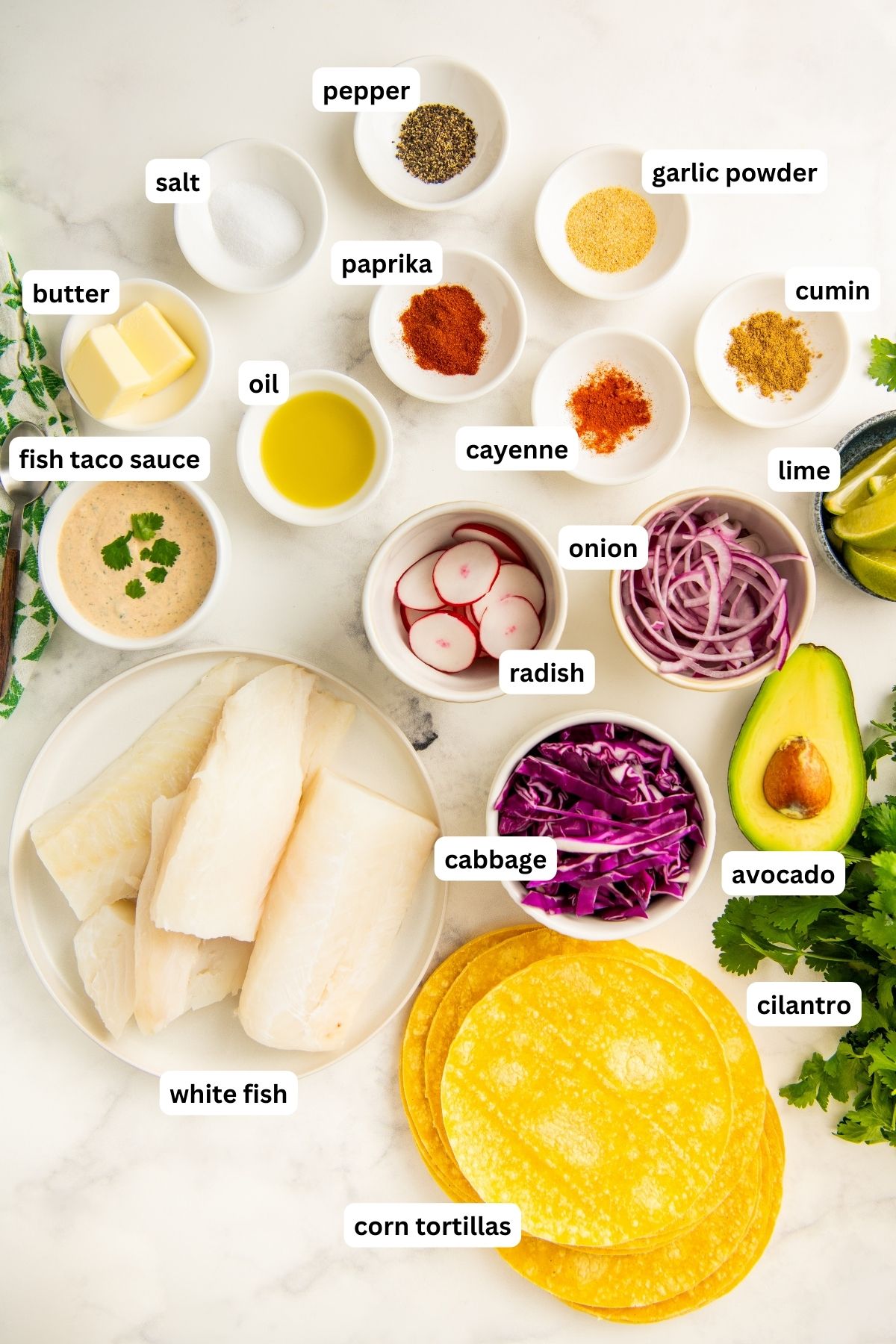 Ingredients for this easy fish tacos recipe arranged in bowls. From top to bottom: pepper, salt, garlic powder, paprika, cumin, cayenne, butter, oil, fish taco sauce, radishes, onion, lime, avocado, cilantro, cabbage, white fish and corn tortillas. 