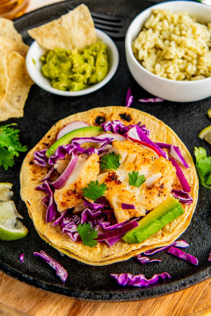 One fish taco being assembled and topped with purple cabbage, fresh cilantro, and avocado.